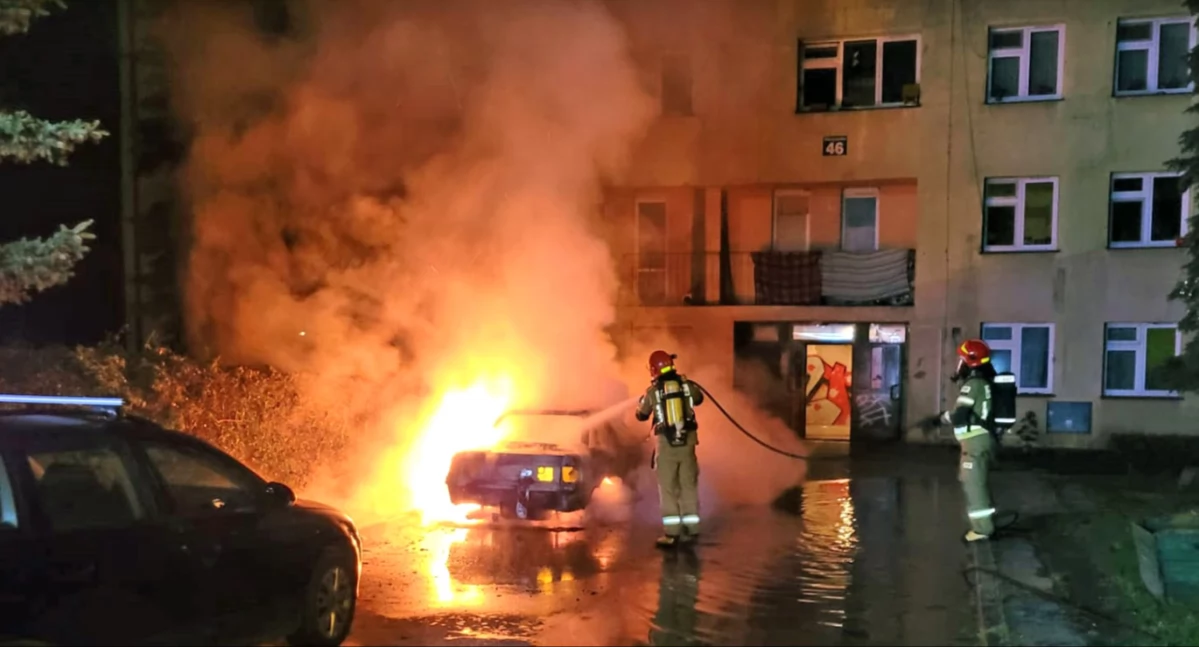 Zdjęcie strażaków, którzy gaszą płonące audi. W blasku płomieni widać budynek mieszkalny