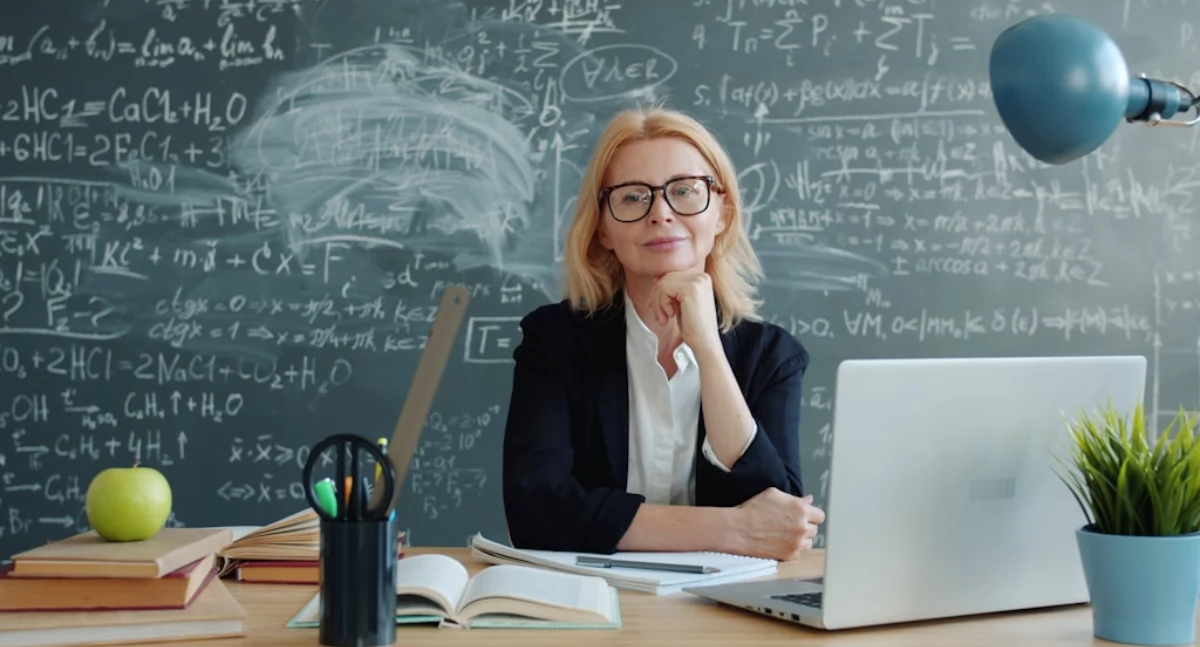 Uśmiechnięta nauczycielka siedząca przy biurku z laptopem, zeszytami i książkami, na tle tablicy zapisanej wzorami i notatkami. Spokojna postawa i przyjazny wyraz twarzy podkreślają wspierające podejście do nauczania dorosłych przygotowujących się do matury po latach. Grafika nawiązuje do kursów maturalnych dla dorosłych w Kielcach. Więcej informacji o ofercie można znaleźć na stronie Symposio.